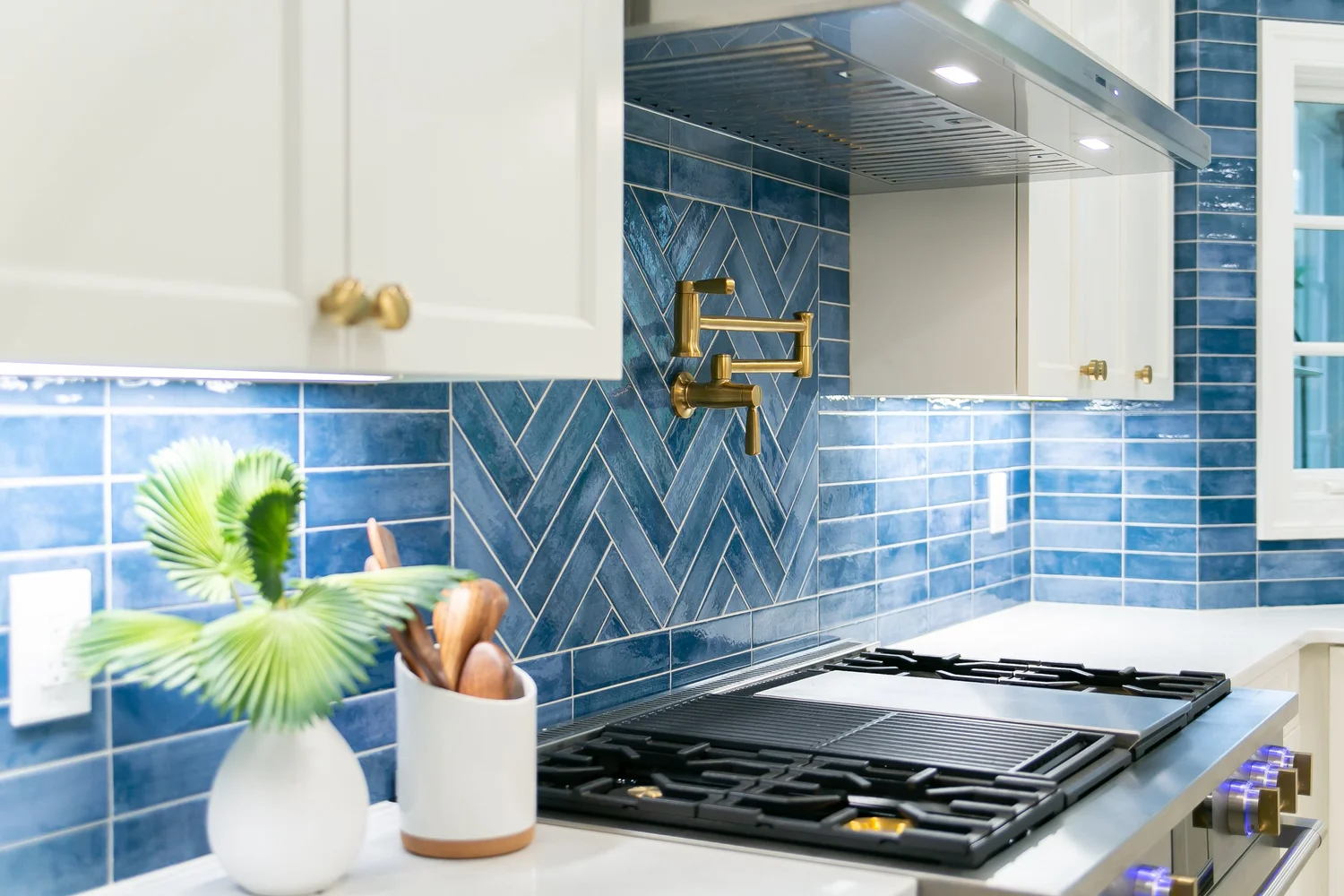 Ford’s Colony kitchen detail featuring a blue herringbone tile backsplash, white cabinetry, brass pot filler, and professional range