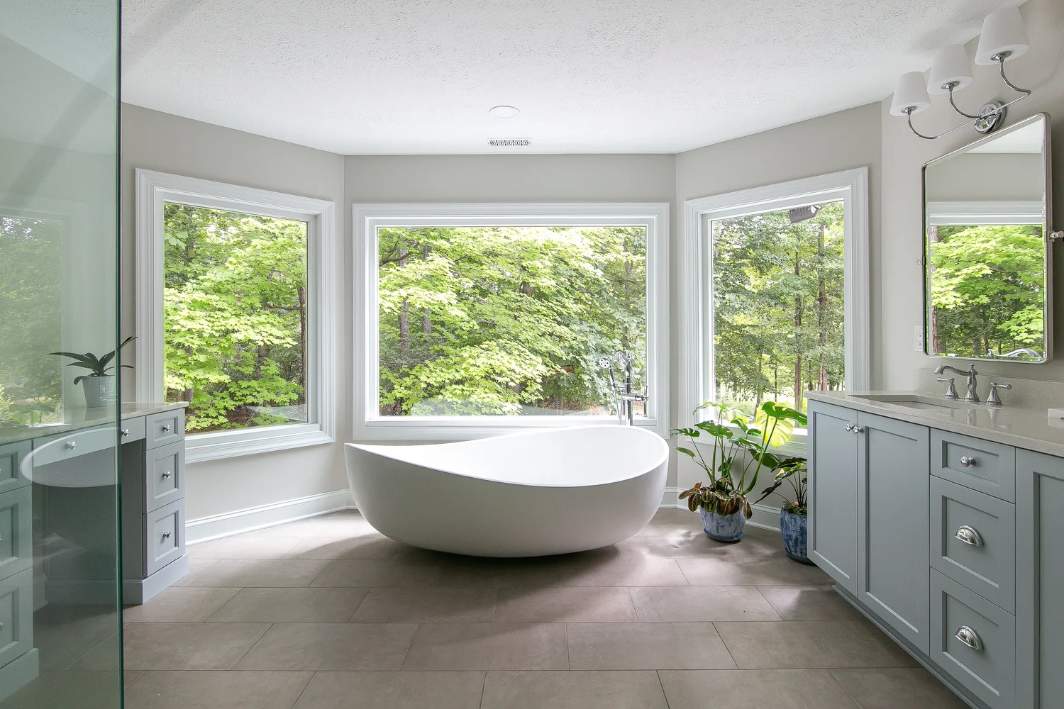 Light-filled Toano primary bathroom with freestanding tub centered beneath three wooded-view windows, dual vanities, and a glass shower