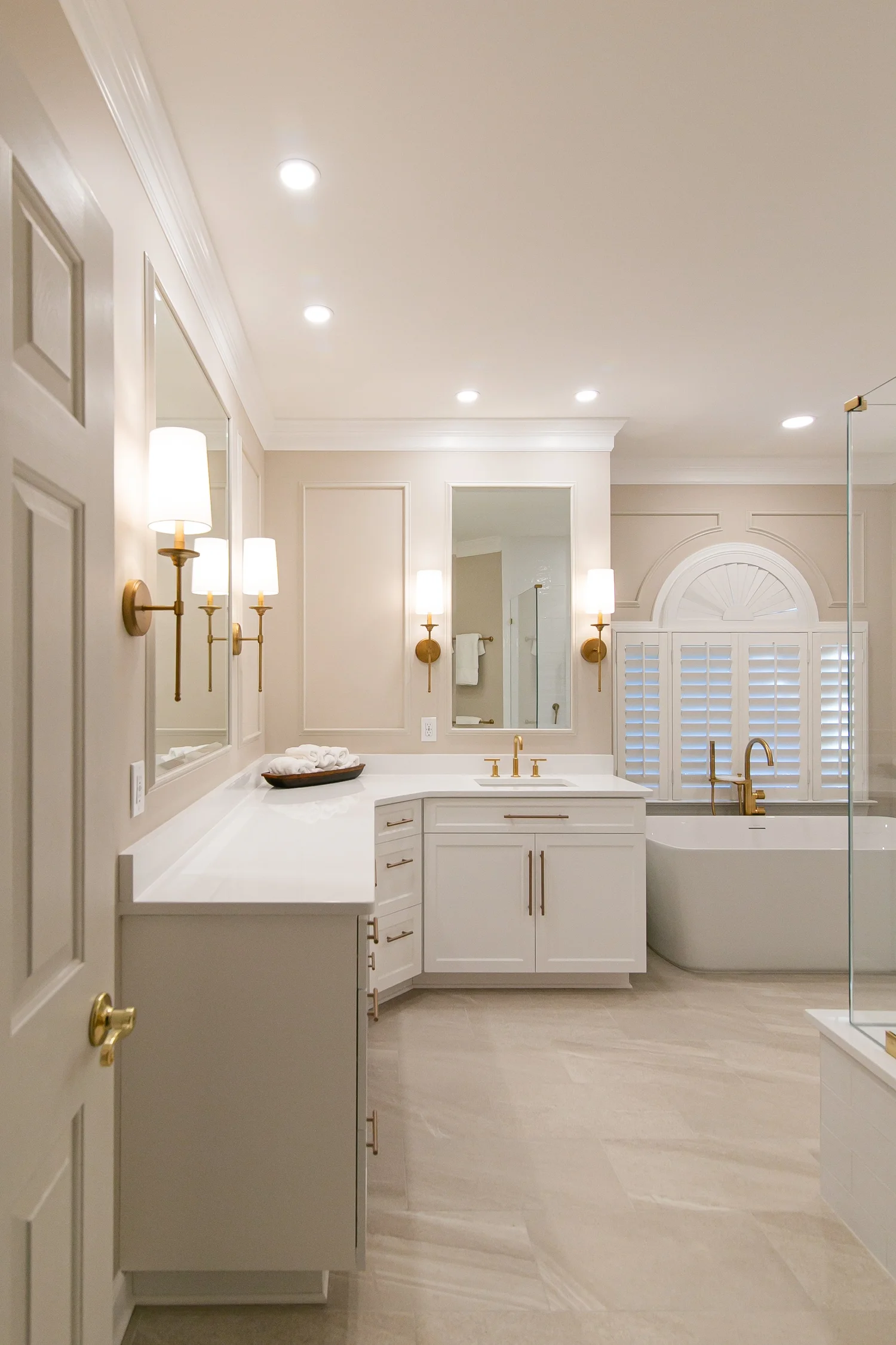 Bright Ford’s Colony primary bathroom with white custom vanities, freestanding tub, frameless glass shower, warm brass fixtures, and layered lighting