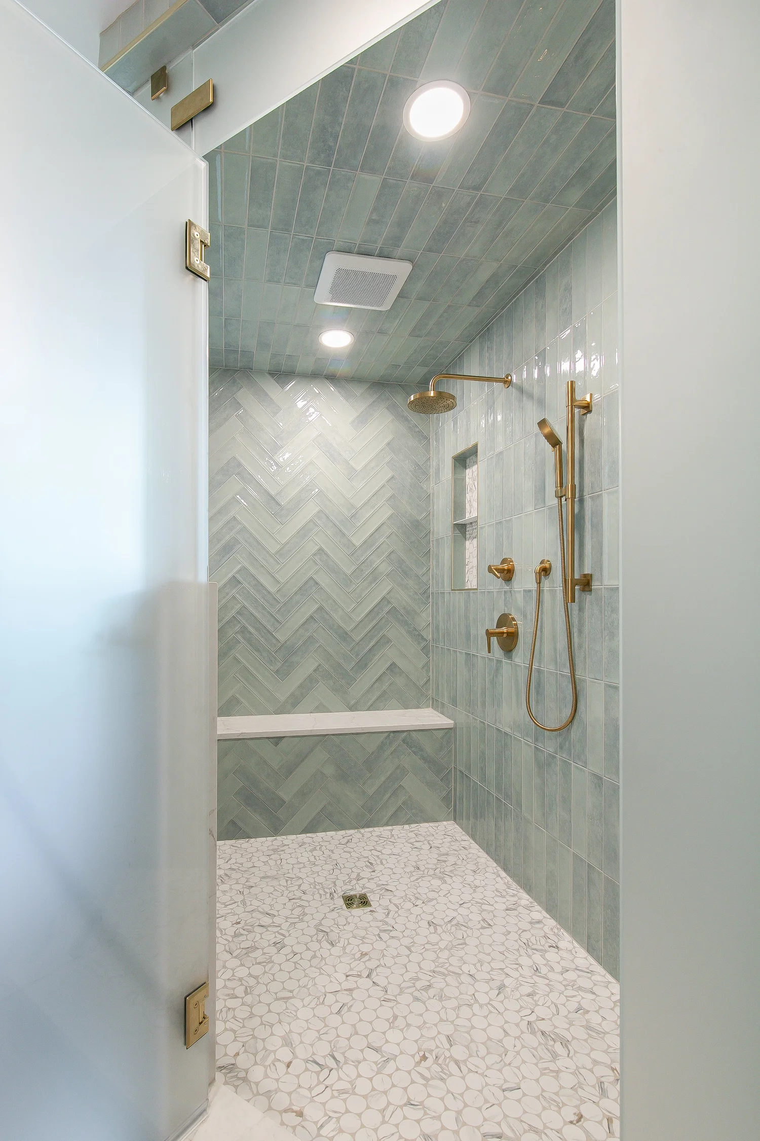 Walk-in shower finished with blue-green tile, herringbone feature wall, pebble mosaic floor, built-in bench, niche, and brass rain shower fixtures