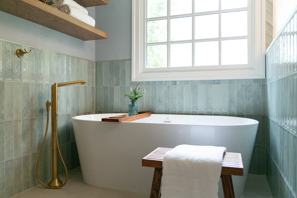 Freestanding white soaking tub with sea-glass blue tile, warm brass floor-mounted filler, natural wood bath tray, and floating shelves in a Ford’s Colony bathroom designed and built by VanFleet Homes