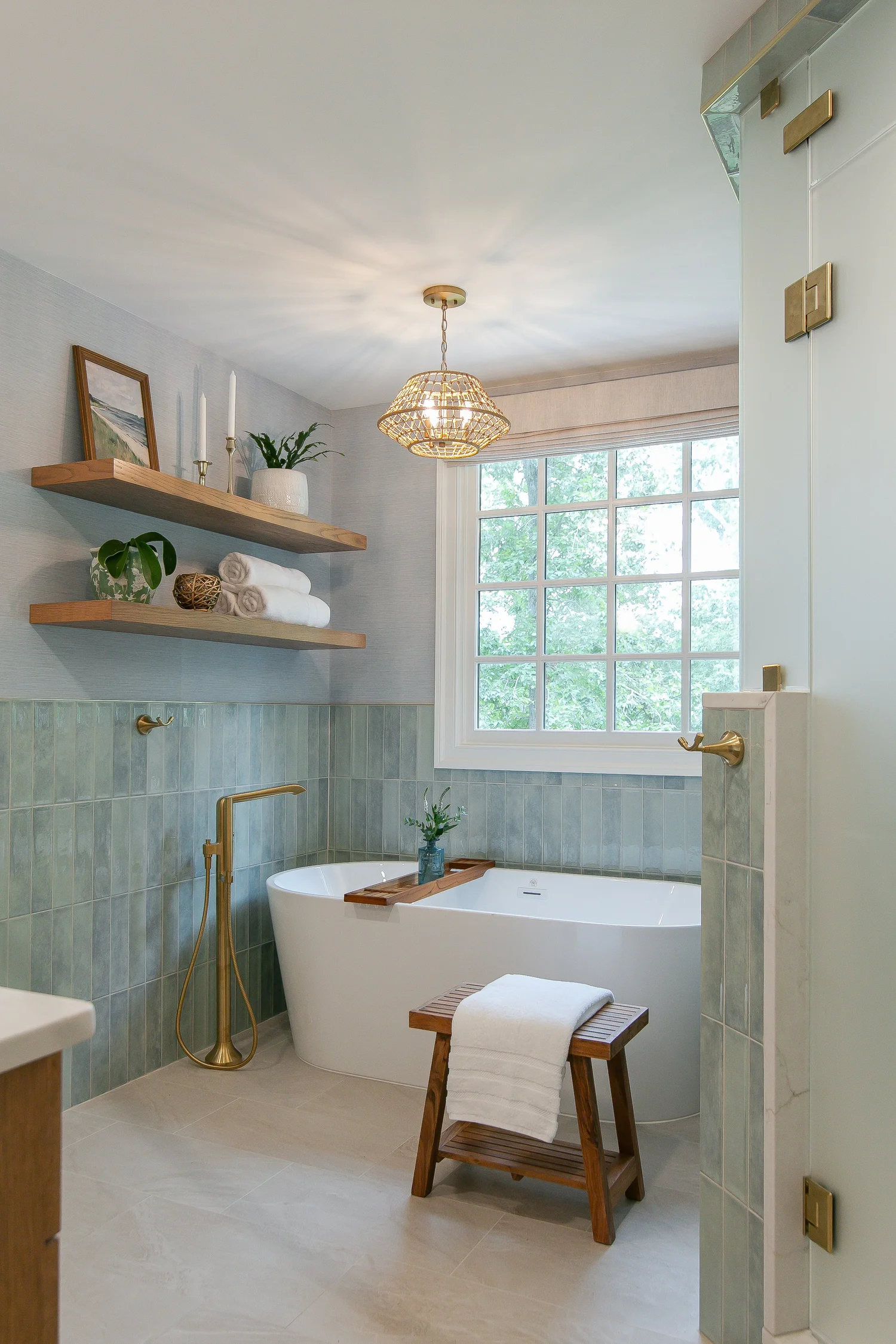 Completed Ford’s Colony bathroom with a freestanding tub, blue-green tile wainscoting, natural wood shelves, woven pendant light, and warm brass fixtures