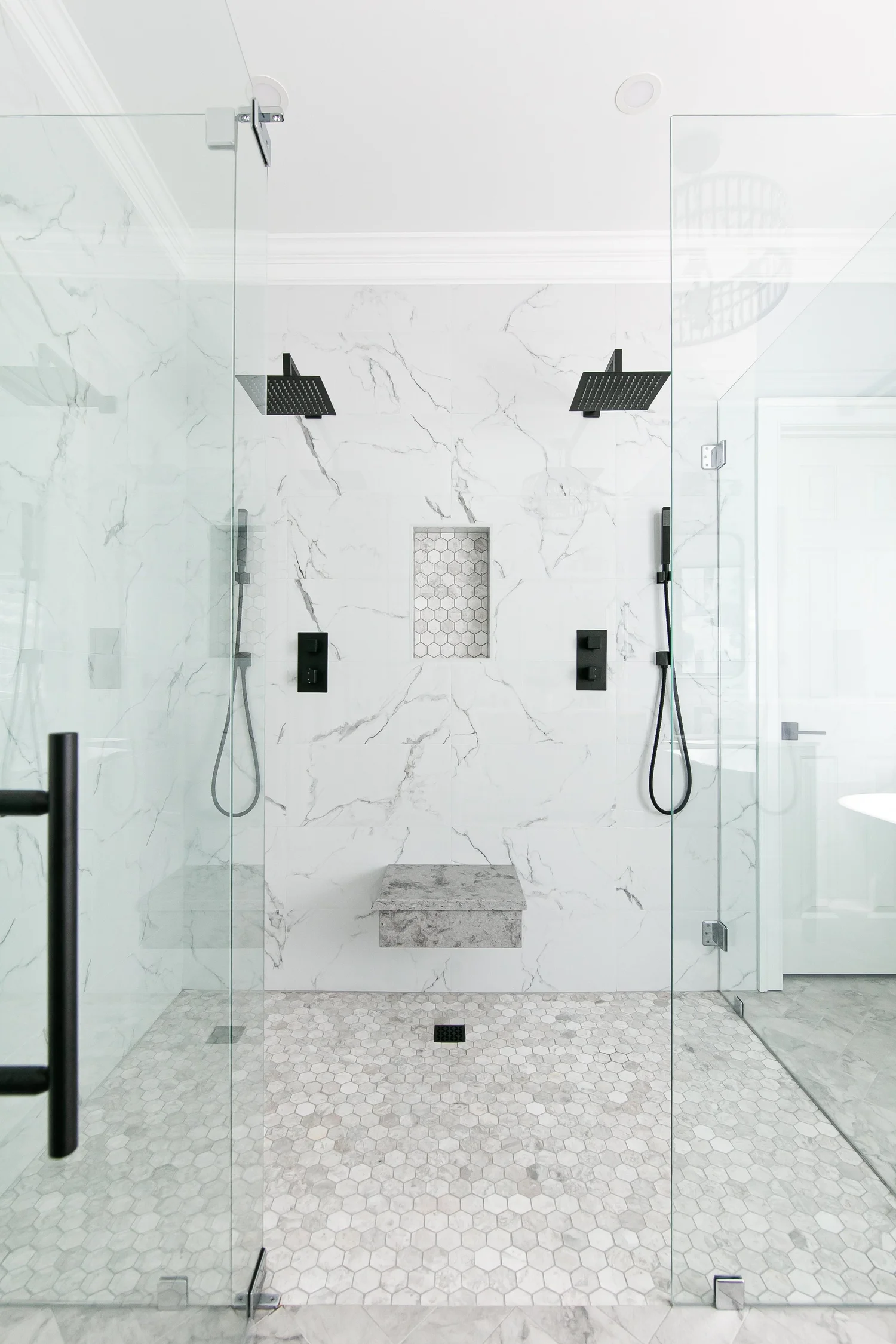 Spacious Greensprings West walk-in shower with marble-look tile, dual black rain heads and handheld fixtures, floating stone bench, niche, and frameless glass